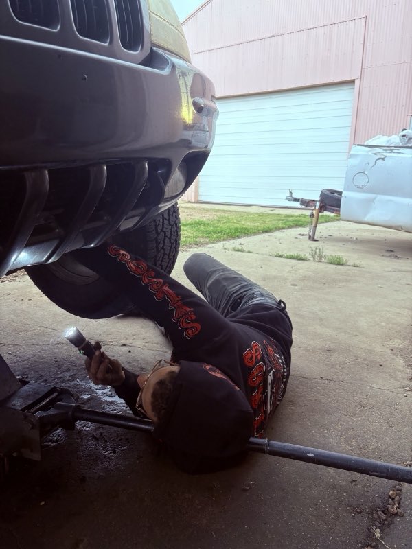 Ryan lying under the front of a vehicle with a flashlight, doing a hands-on repair