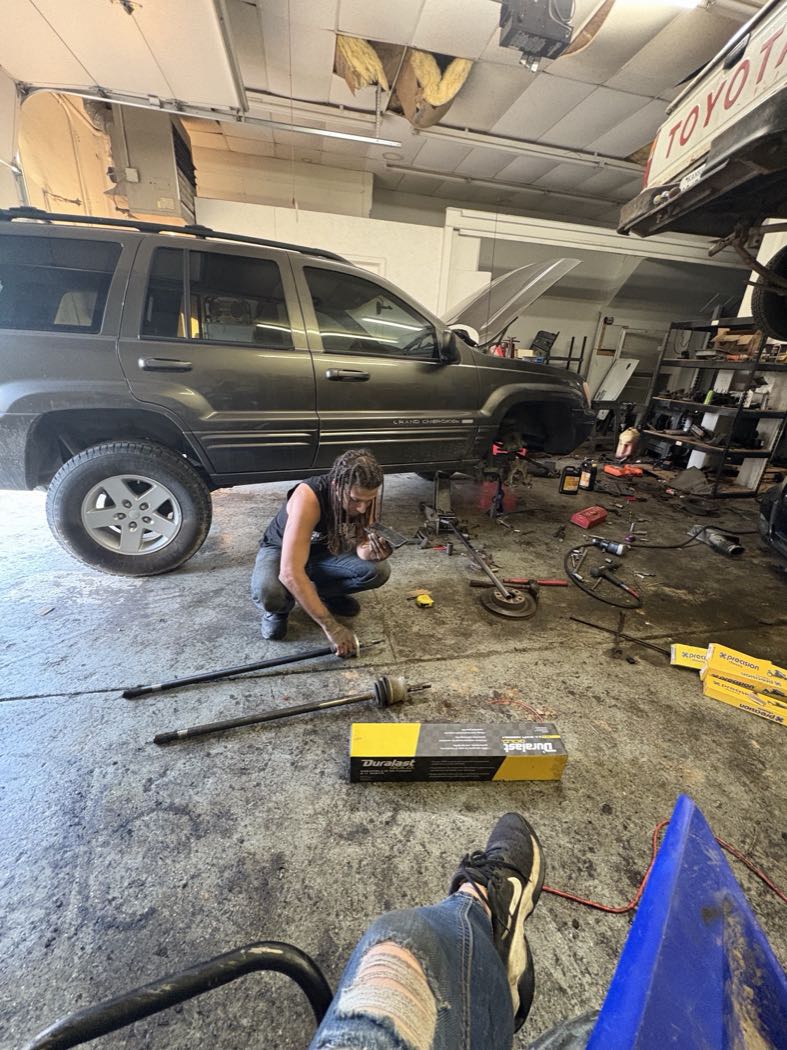 Ryan crouched in the shop bay working on a drivetrain repair, with new parts laid out beside a Jeep Grand Cherokee